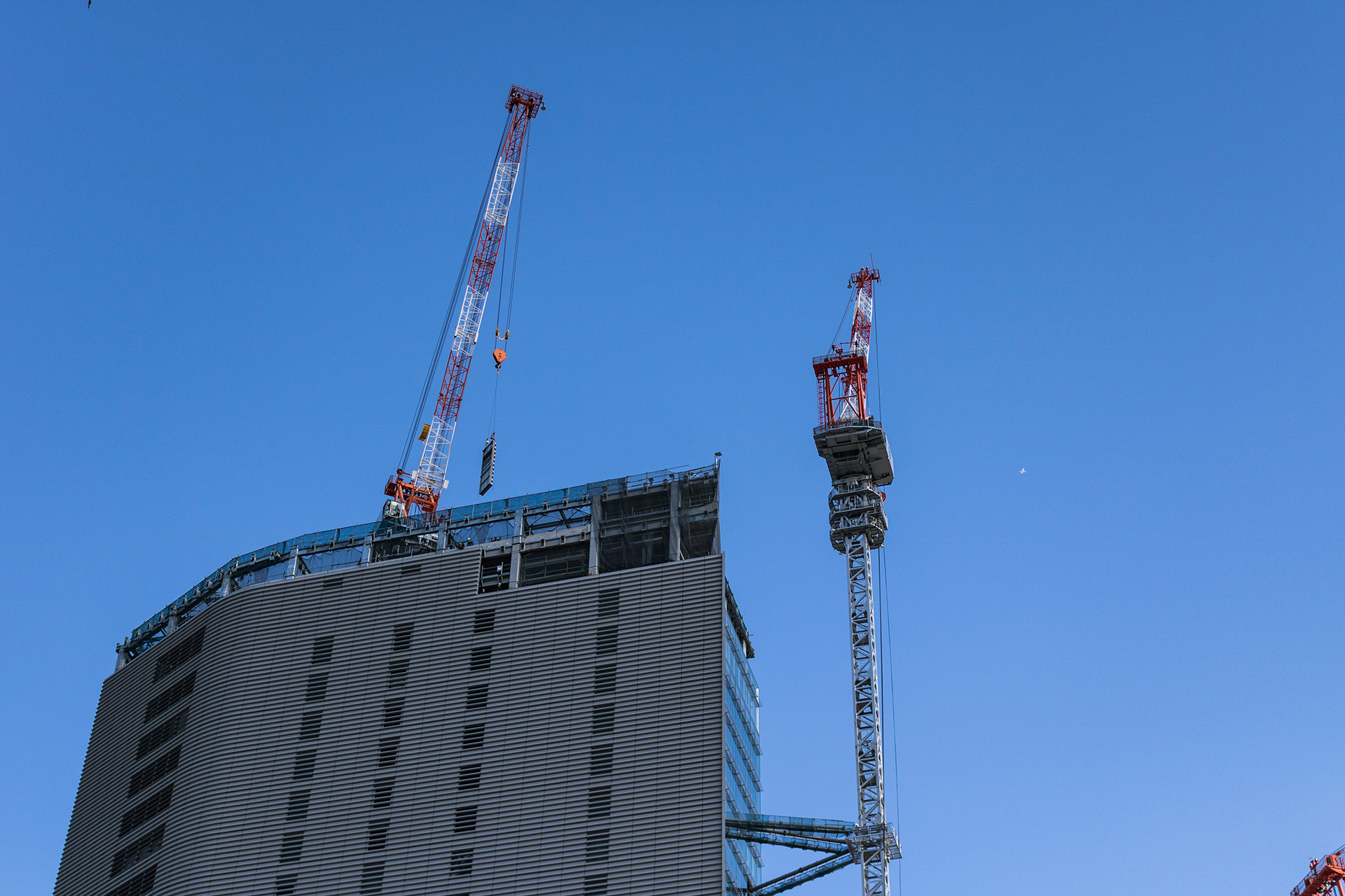 Two red and white cranes on top of and next to a modern skyscraper with the blue sky behind them.